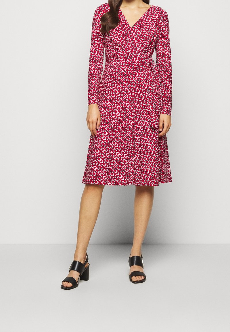 Czerwona sukienka w wzory z długim rękawem, dekoltem w kształcie V i rozkloszowanym dołem. Posiada geometryczny wzór w białym kolorze. Połączona z czarnymi sandałami na słupku.