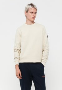 Cream sweatshirt with a round neckline and ribbed cuffs, paired with dark pants featuring red text, set against a solid white background.