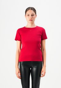 Woman wearing a red short-sleeve T-shirt with "HUGO" logo and black leather pants, standing against a plain white background.