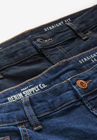 Donkerblauwe jeans met rechte pasvorm en lichtbruine contrasterende stiksels, met een knoopsluiting en labels voor maat en merk.