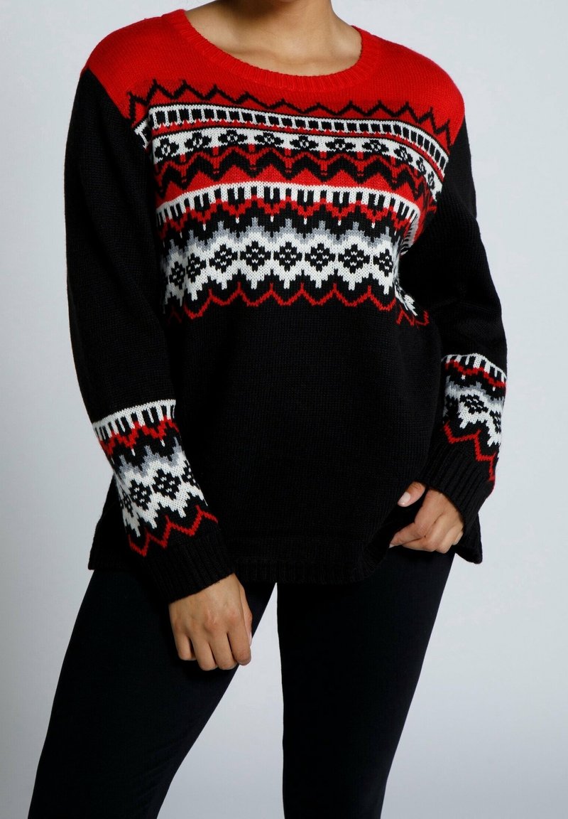 Personne portant un pull en maille à motifs noirs, rouges et blancs avec des motifs en zigzag et floraux, associé à un pantalon noir, debout dans une posture détendue.