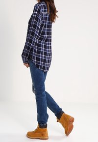 Person wearing blue plaid shirt, blue jeans, and tan lace-up boots, standing with one leg slightly raised against a plain background.