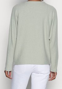 Ljusgrön ribbad stickad tröja med rundad halsringning och långa ärmar, tillsammans med vita jeans, sedd bakifrån.