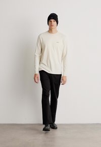 Cămașă crem cu mânecă lungă și un logo subtil, pantaloni negri slim-fit și o căciulă neagră; material mai fin cu un design simplu și curat.