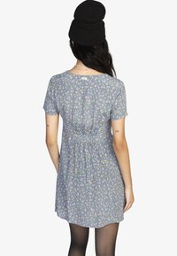 Himmelblaues Kleid mit einem Blumenmuster in Pink und Gelb, kurzen Ärmeln, einem gerafften Bund und einem runden Rückenausschnitt. Mit einem Anhänger akzentuiert.