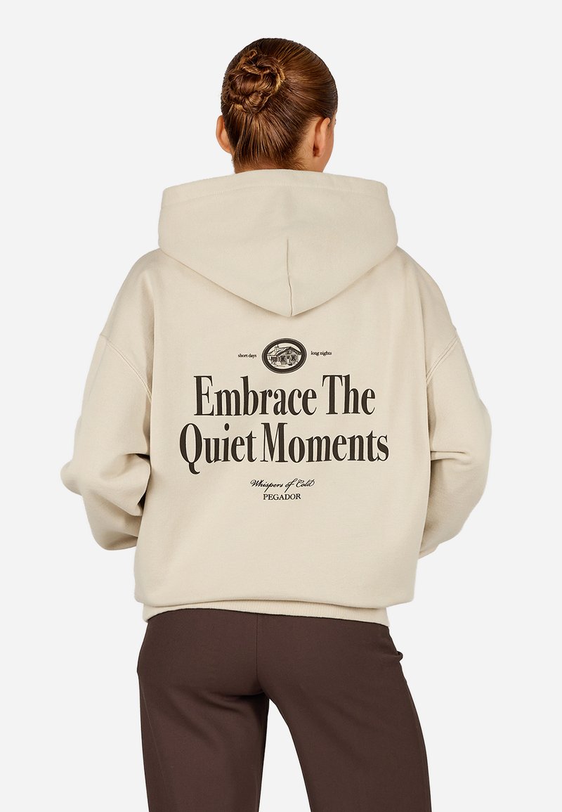 Bežová oversized mikina s veľkým čiernym potlačou na zadnej strane. Materiál vyzerá mäkký, s priestrannou kapucňou a rebrovanými manžetami.