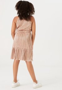 Růžovo-zlaté plisované šaty s rozšířeným lemem a halter dekoltem. Kombinované s bílými teniskami, které zdůrazňují hladkou strukturu a lesklý povrch.