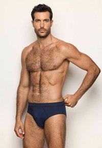 Hombre musculoso con vello en el pecho y los brazos, vistiendo unos calzoncillos azul marino, ajustándose la cinturilla, contra un fondo claro y liso.