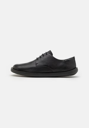 Zapatos de cuero negro con un acabado suave, punta redondeada y un mínimo de cordones. Presentan una suela de goma plana y una marca sutil en el costado.