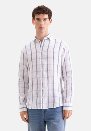Mand iført en hvid skjorte med blå lodrette og vandrette striber og blå jeans, der står mod en ensfarvet baggrund.