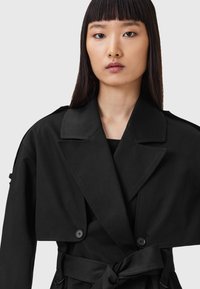 Giovane donna con lunghi capelli neri lisci che indossa un trench coat nero con cintura, dotato di un ampio sovrapposto corto e reverse.
