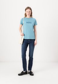 Šviesiai mėlynas medvilninis marškinėlis su žodžiu "BOSS" tamsiai mėlyna spalva, derinamas su tamsiai džinsiniais džinsais ir juodais loafer'iais. Modelis laiko kreminės spalvos maišą.