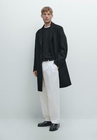 Lungo cappotto nero, maglione nero e pantaloni bianchi. Scarpe nere completano l'outfit. Design semplice e strutturato con un contrasto di colori pulito.