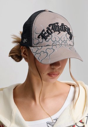 Kobieta nosząca beżową i czarną czapkę typu trucker z napisem "Ed Hardy" i wzorami serc, z włosami związanymi do tyłu i dwoma luźnymi kosmykami przy twarzy.
