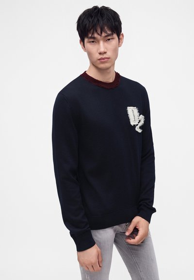 Jeune homme aux cheveux noirs portant un pull noir avec un col rouge et un logo blanc texturé, ainsi qu’un jean gris clair, debout devant un fond blanc.