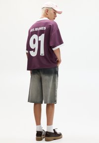 Oversized purple jersey with white accents, "LES JEUNES" and "91" on the back. Paired with gray denim shorts and brown shoes.