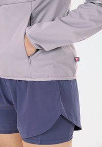 Person iført grå jakke med hånden i lynlåslommen og mørkeblå lagdelte sportshorts synlige mod hvid baggrund.
