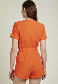 Combinaison en coton orange avec des manches courtes, un tissu texturé, des poignets roulés et une ceinture à nouer à la taille, présentant une coupe décontractée.