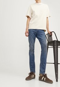 Camiseta de gran tamaño color beige, jeans ajustados azules, zapatillas de ante marrón con rayas blancas. Hombre de pie junto a una silla de metal perforado negra.