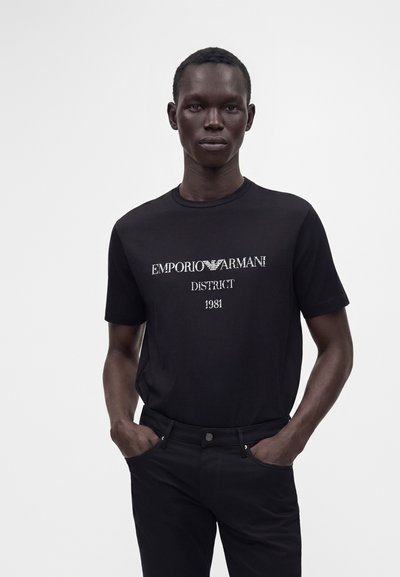 Άνδρας που φοράει μαύρο t-shirt Emporio Armani με λευκό κείμενο και μαύρο παντελόνι, στέκεται με τα χέρια στις τσέπες σε απλό φόντο.