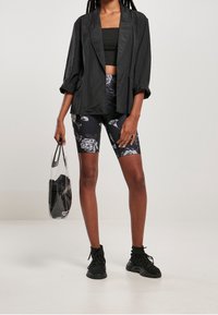 Kvinna klädd i en svart croppad topp, svart blazer, blommiga cykelshorts, svarta sneakers, som håller i en genomskinlig axelväska med svarta föremål inuti.