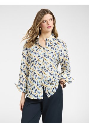 Kobieta w niebiesko-żółtej koszuli na guziki z wzorem, z podwiniętymi rękawami i ciemnymi spodniami, patrząca nieco na bok.