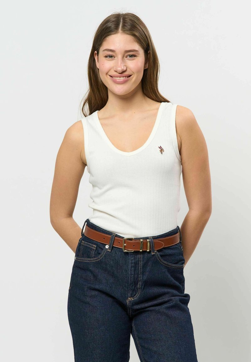 Jeune femme souriante, portant un haut blanc sans manches avec un petit logo et un jean bleu foncé avec une ceinture marron, debout devant un fond uni.