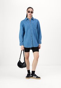 Denim srajca v svetlo modri barvi z gumbom spredaj in žepom, v kombinaciji s črnimi kolesarskimi hlačami in črnimi supergami. Mala črna torbica in sončna očala.
