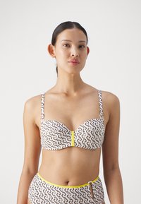 Top bikini in tessuto bianco con forme marroni intrecciate, dotato di un accento centrale giallo e spalline sottili regolabili.