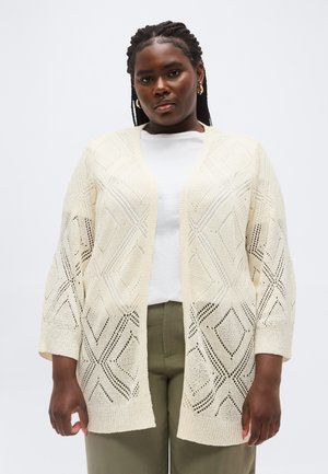 Γυναίκα που φοράει ανοιχτό μπροστά εκρού πλεκτή ζακέτα με μοτίβα ρόμβου, λευκό πουκάμισο και παντελόνι ελιάς, που στέκεται μπροστά σε απλό φόντο.