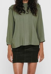 Vrouw in een losse olijfgroen blouse met lange mouwen en een donkergroen corduroy minirok, staande tegen een effen achtergrond.