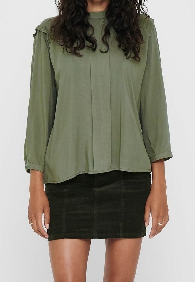 Vrouw in een losse olijfgroen blouse met lange mouwen en een donkergroen corduroy minirok, staande tegen een effen achtergrond.