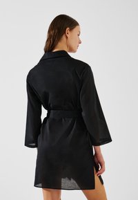 Toga negra hecha de tela ligera, con cuello, mangas largas, cintura con cinturón y aberturas laterales para facilitar el movimiento.