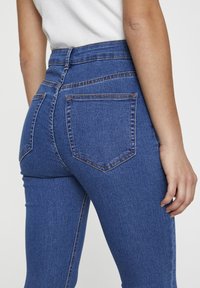 Personne portant un jean en denim bleu taille haute et un haut blanc, vue de dos, une main reposant sur le côté.