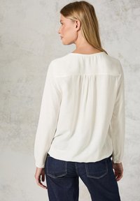 Lättviktig vit blus med långa ärmar, subtil texturerad mönster och en rundad nederkant. Har en ryggsöm och sammanknuten detalj.