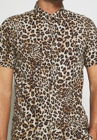 Mand iført skjorte med korte ærmer og knapper, prydet med brun, sort og beige leopardmønster mod en ensfarvet baggrund.
