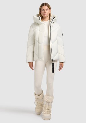 Balts pūkains mētelis ar augstu kaklu, dimantu formas izšuvumu un rāvējslēdzēju, kombinēts ar krēmkrāsas legingiem un teksturētām krēmkrāsas zālēm.