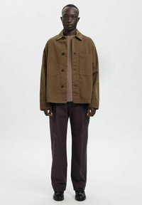 Brązowa bawełniana kurtka zapinana na guziki z bocznymi kieszeniami i luźnym krojem, zestawiona z ciemnofioletowymi, prostymi spodniami oraz czarnymi butami.