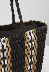 Torebka tote wykonana z plecionki z czarnymi, białymi i beżowymi wzorami, solidnymi uchwytami oraz zamykaną na zamek czarną tkaniną w środku.