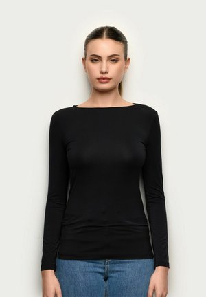 Kobieta z prostymi włosami związanymi w kucyk, ubrana w dopasowaną czarną bluzkę z długim rękawem i niebieskie dżinsy, stojąca na jasnym, jednolitym tle.