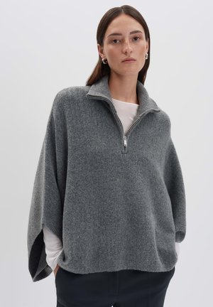 Woman with straight brown hair wearing silver hoop earrings, a gray zip-up wool poncho, white long-sleeve top, and black pants on white background.