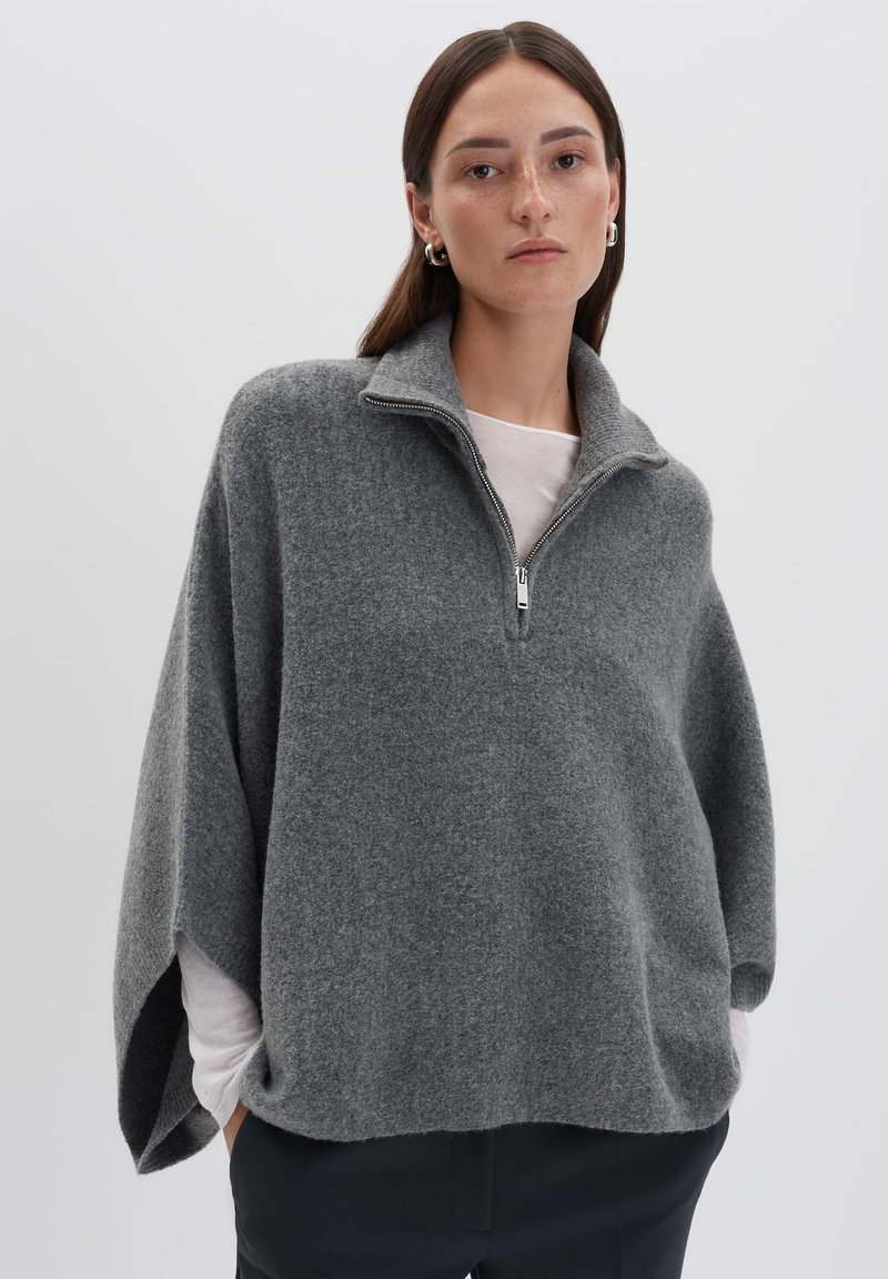 Frau mit glattem braunem Haar, die silberne Creolen trägt, einen grauen Wollponcho mit Reißverschluss, ein weißes Langarmshirt und schwarze Hosen vor weißem Hintergrund.