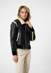Zwarte leren jas met beige schapenvacht kraag en afwerking, voorzien van een knoopsluiting en geribde manchetten. Soepele textuur met contrasterende accenten.