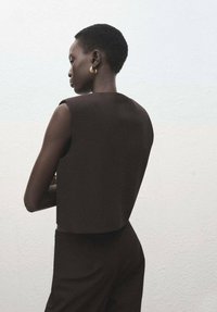 Bruin mouwloos topje met een gestructureerd ontwerp. Gladde stof, cropped lengte, gecombineerd met bijpassende bruine onderkleding. Gouden creolen benadrukken de look.