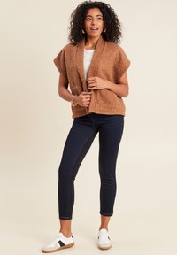 Brauner gestrickter Cardigan mit kurzen Ärmeln, strukturiertem Oberflächen, vorderen Taschen, kombiniert mit dunklen Jeans und weißen Sneakern mit schwarzen Akzenten.