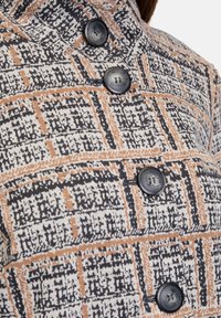 Blazer med ett rutigt mönster i vitt, svart och orange nyanser. Har stora svarta knappar och en strukturerad kragdesign.