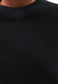 Detail osoby nosící černý svetr s texturovaným vzorem a kulatým výstřihem.