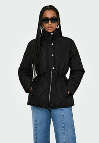 Veste matelassée noire avec col montant, fermeture éclair et taille élastique. Présente des boutons-pression et des poches latérales. Portée sur un jean large bleu.