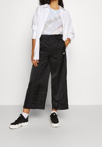 Kvinna som bär svarta vida capribyxor, en tie-dye-t-shirt, en vit öppen långärmad skjorta, svarta och vita sneakers, står med ena handen i fickan.