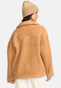 Giacca oversize in finta pelliccia shearling color beige con colletto, tasche laterali e un piccolo dettaglio dorato sul retro. Abbinata a un berretto lavorato a maglia di colore marrone chiaro.